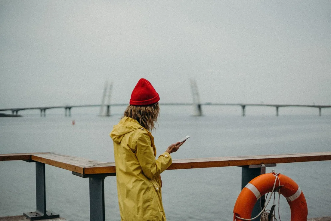 Woman in a yellow raincoat looking at her phone by the water